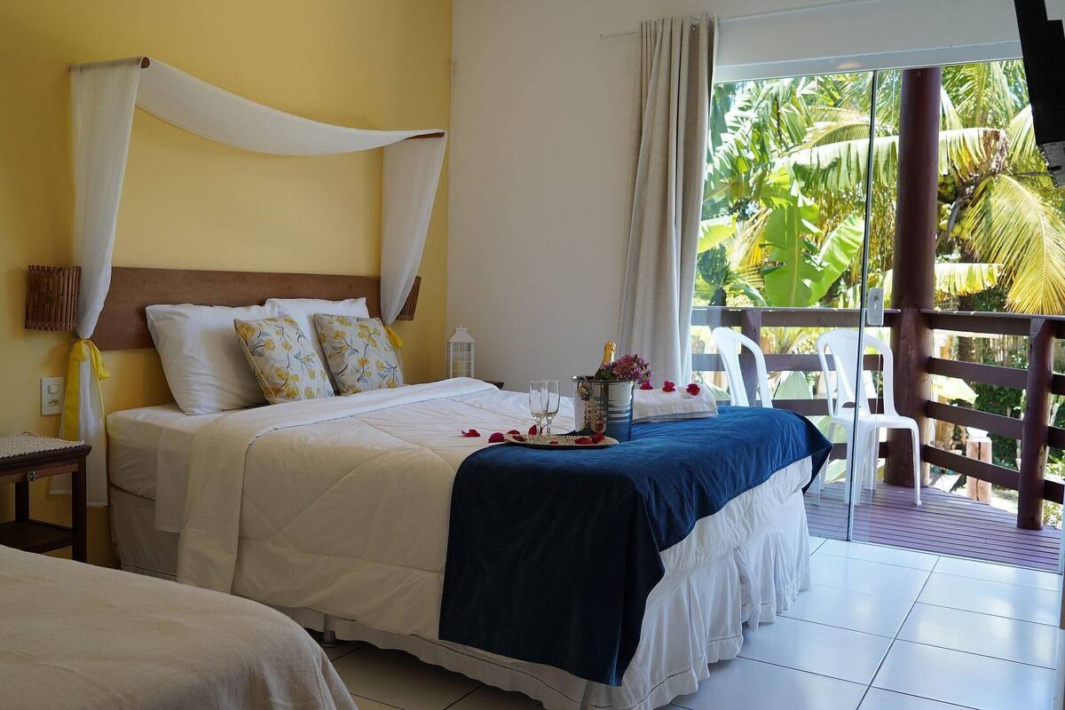 A hotel room with a bed, a nightstand, a bedside table, a window with a view of palm trees, and a balcony.
