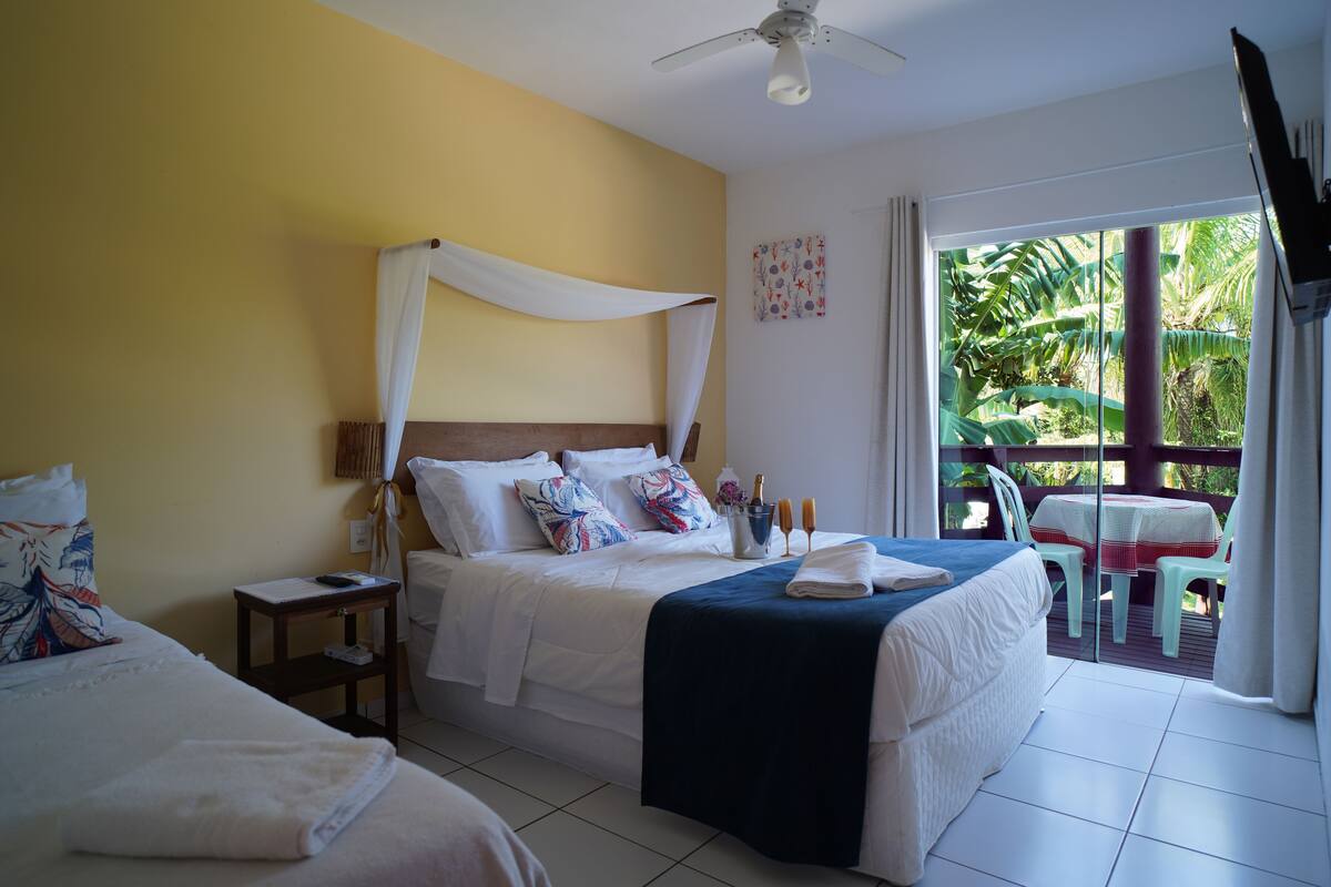 A hotel room with two beds, a ceiling fan, a balcony with a table and chairs, and a view of greenery outside.