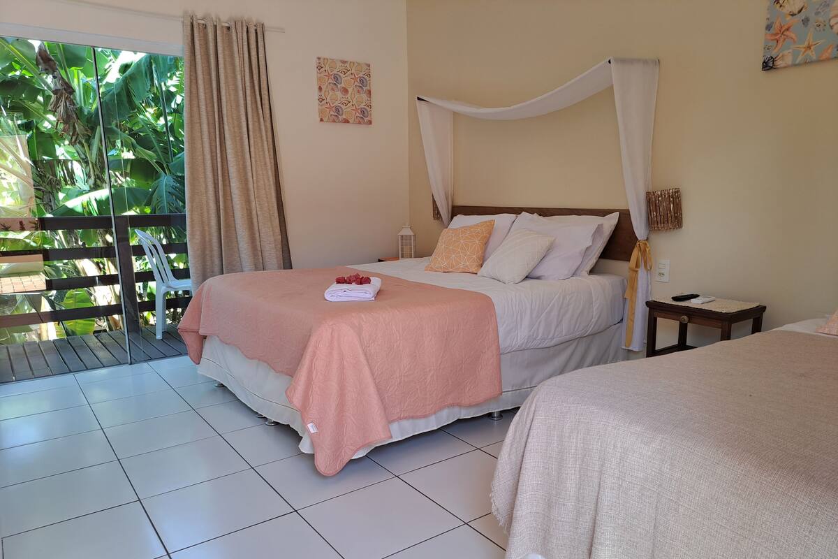 A hotel room with a bed, a nightstand, a small table, and a view of a balcony with plants.