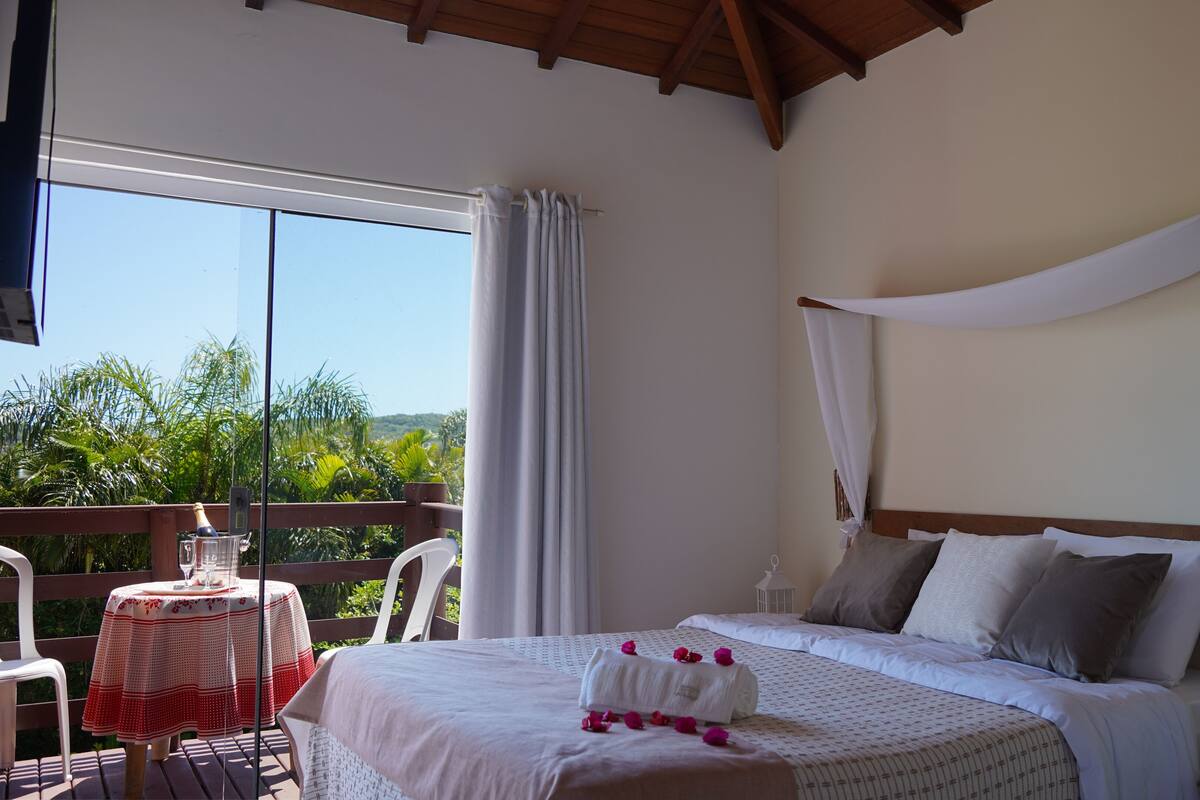 A bedroom with a bed, a bedside table, a nightstand, a window with curtains, and a view of greenery outside.
