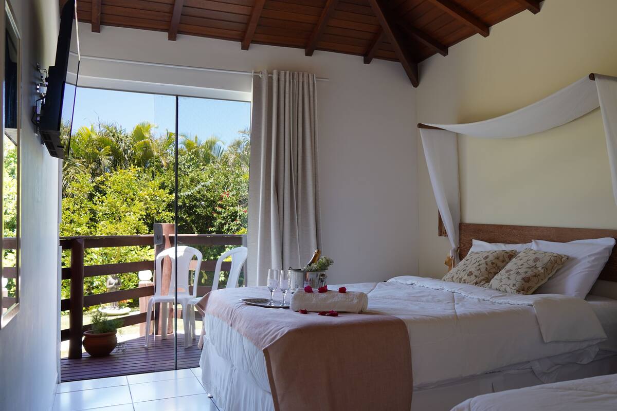 A bedroom with a bed, a bedside table, a nightstand, a window with curtains, and a view of greenery outside.