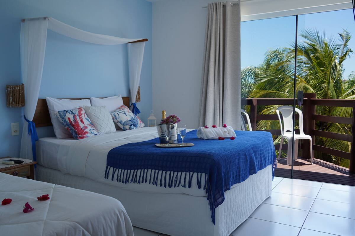 A hotel room with a large bed, a smaller bed, a balcony with a view of palm trees, and a minibar with a bottle of champagne and flowers.