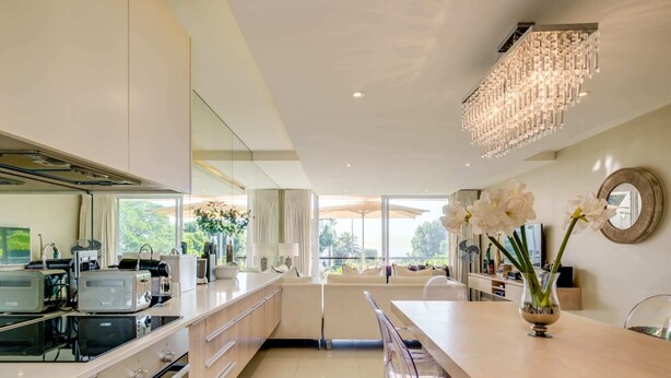 A modern kitchen with a central island, a dining area with bar stools, and a large window offering a view of greenery.