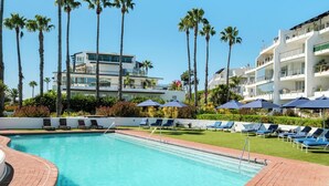A swimming pool with lounge chairs and umbrellas, surrounded by palm trees and modern buildings.