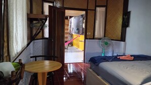 A small, compact room with a bed, a table, and a chair. There is a fan on the bed and a window with curtains. - Koh Chang Garden Lodge Guest  Room G (Klong Son)