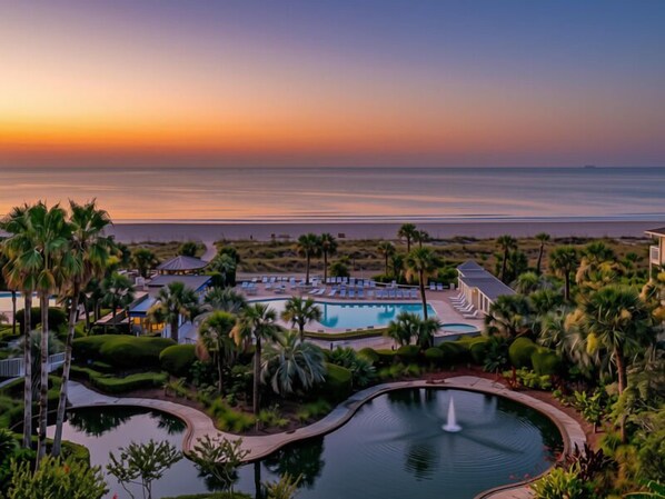 A resort with a pool, palm trees, and a view of the ocean at sunset.