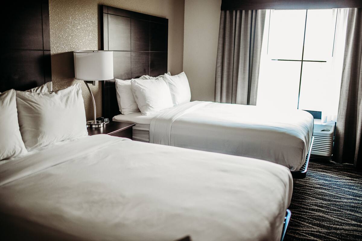 A hotel room with two beds, a nightstand, a lamp, and a window with curtains.