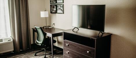 Une chambre d’hôtel équipée d’un bureau, d’une chaise, d’une télévision, d’une commode et d’une fenêtre avec des rideaux.