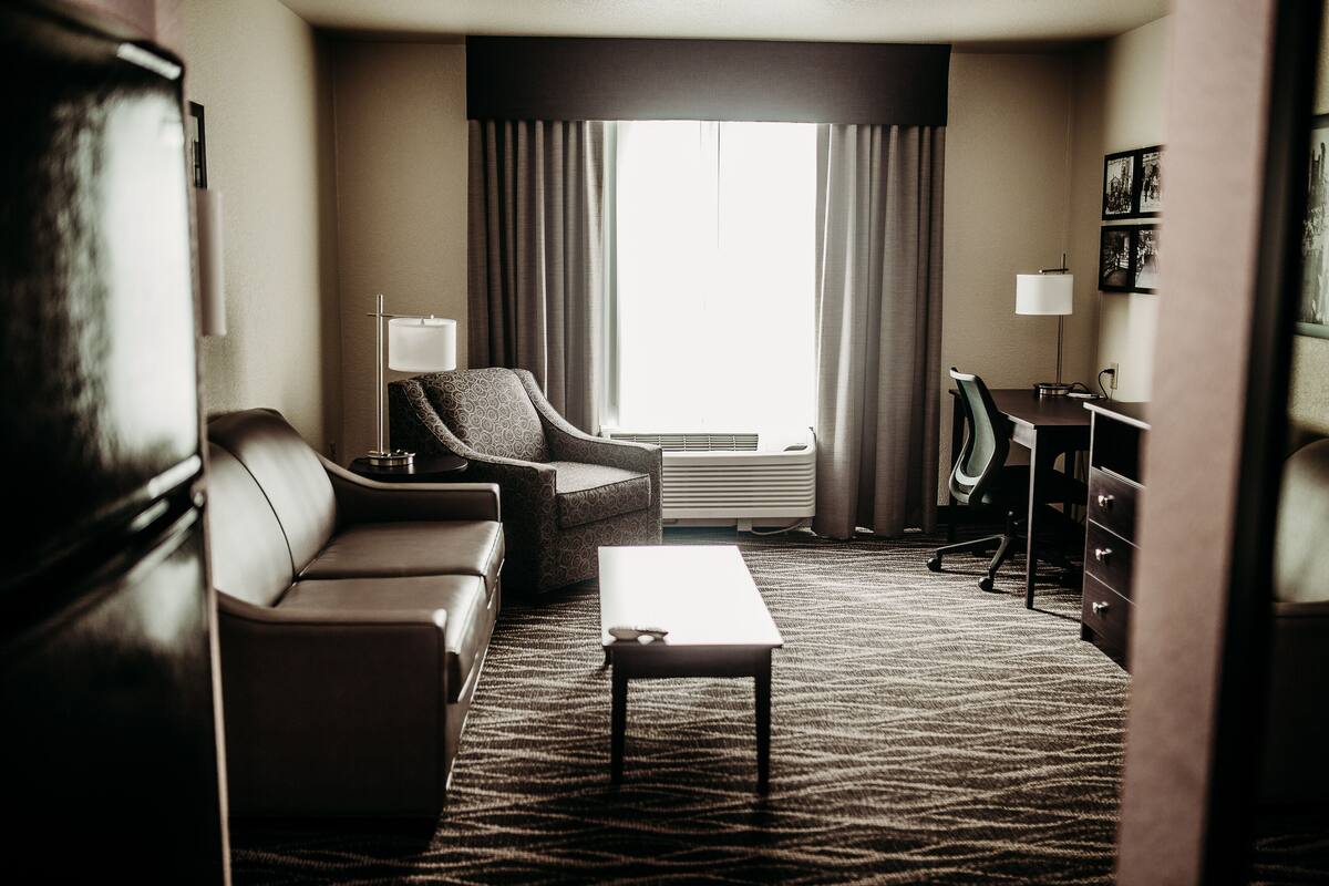 A hotel room with a leather sofa, a small table, a desk with a chair, and a large window with curtains.