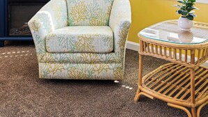 Ein Zimmer mit gelben Wänden, einem großen Flachbildfernseher, einem gemusterten Sessel, einem kleinen Korbstuhl mit einer Pflanze und einem Fenster mit Jalousien.