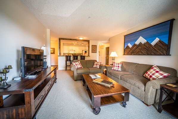 A living room with a TV, sofa, coffee table, and a painting of mountains. - 2-Bedroom 2-Bathroom Condo with Mountain Views (Radium Hotsprings)