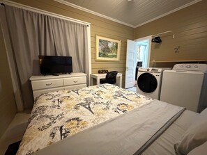 Habitación de hotel con cama, televisión, escritorio, silla, lavadora y puerta que da a otra habitación.