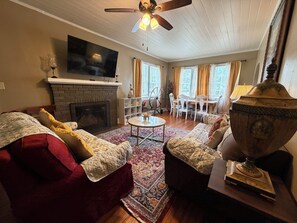 A living room with a fireplace, a television mounted above it, a ceiling fan, and a dining area with a table and chairs. - Spacious 4BR  Sleeps 12+ VSU, SGMC + Parking (Valdosta)