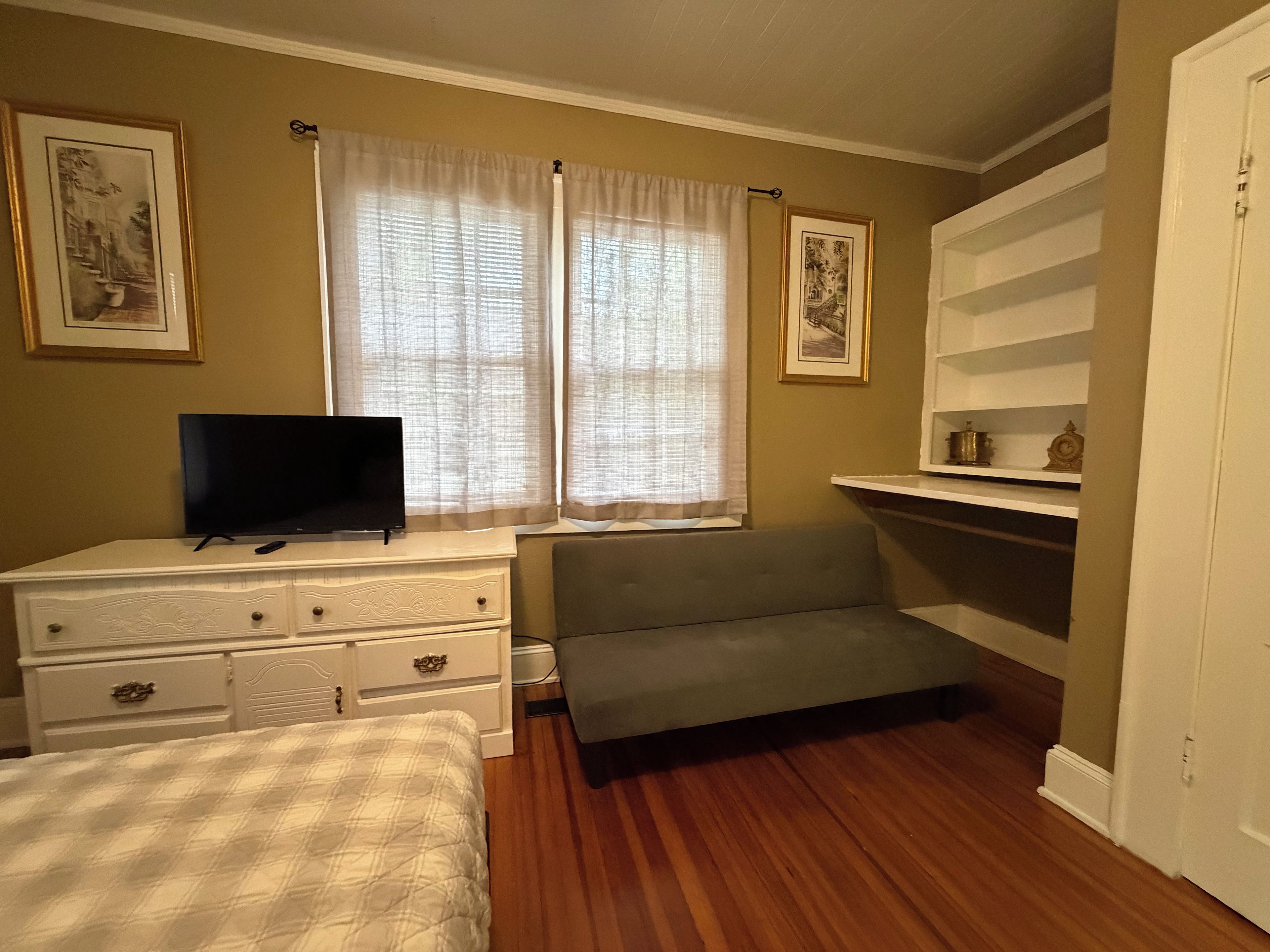 Une chambre à coucher avec un lit, une commode, une télévision et une fenêtre avec des rideaux.