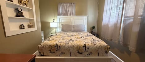 A bedroom with a bed, a nightstand, a lamp, a shelf with decorative items, and a window with curtains.