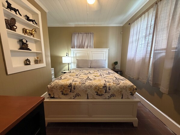 A bedroom with a bed, a nightstand, a lamp, a shelf with decorative items, and a window with curtains.