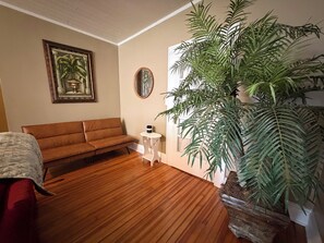 A room with a wooden floor, a brown sofa, a small white table, a large potted plant, and a framed picture on the wall.