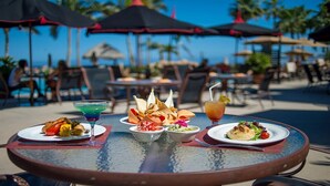 A table set with various dishes and drinks at a beachside restaurant with umbrellas and palm trees in the background.
