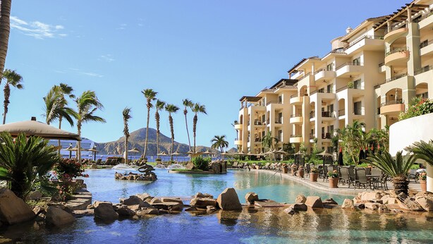A resort pool with palm trees, a mountain in the background, and a multi-story building with balconies.
