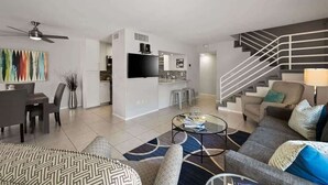 Un salon moderne avec un canapé, une table basse et un tél éviseur. On aperçoit un escalier et un coin cuisine au fond de la pièce.