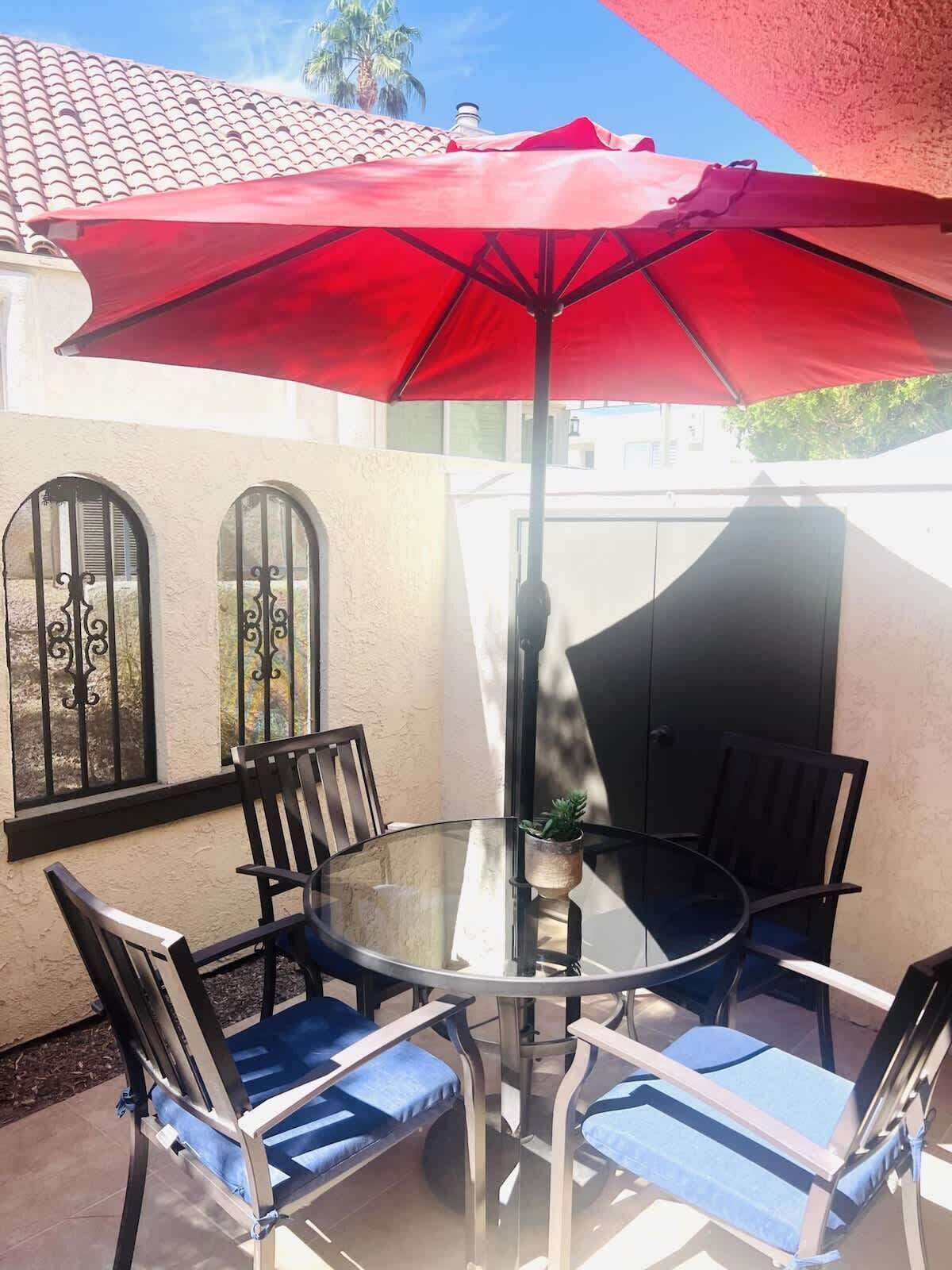 Une table ronde en verre, quatre chaises, un parasol rouge et une petite plante en pot.