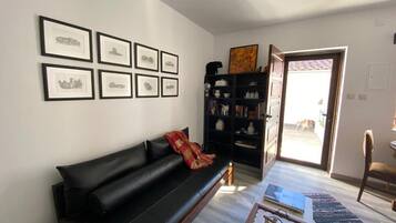 Un salon avec un canapé en cuir noir, une bibliothèque, des tableaux encadrés accrochés au mur et une vue sur l’extérieur par une porte ouverte.