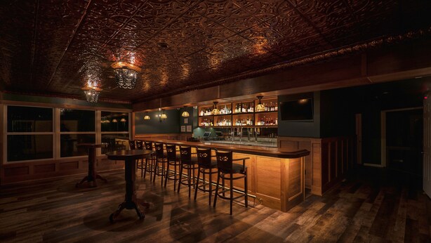 A bar area with wooden bar stools, a bar counter, and a ceiling with intricate designs. - The Inn of Lake George (Lake George)