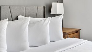 A neatly made bed with white linens and pillows, a grey upholstered headboard, and a wooden nightstand with a white lamp. - The Inn of Lake George (Lake George)