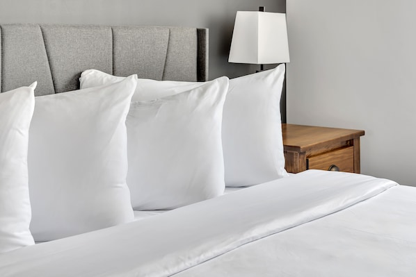 A neatly made bed with white linens and pillows, a grey upholstered headboard, and a wooden nightstand with a white lamp.