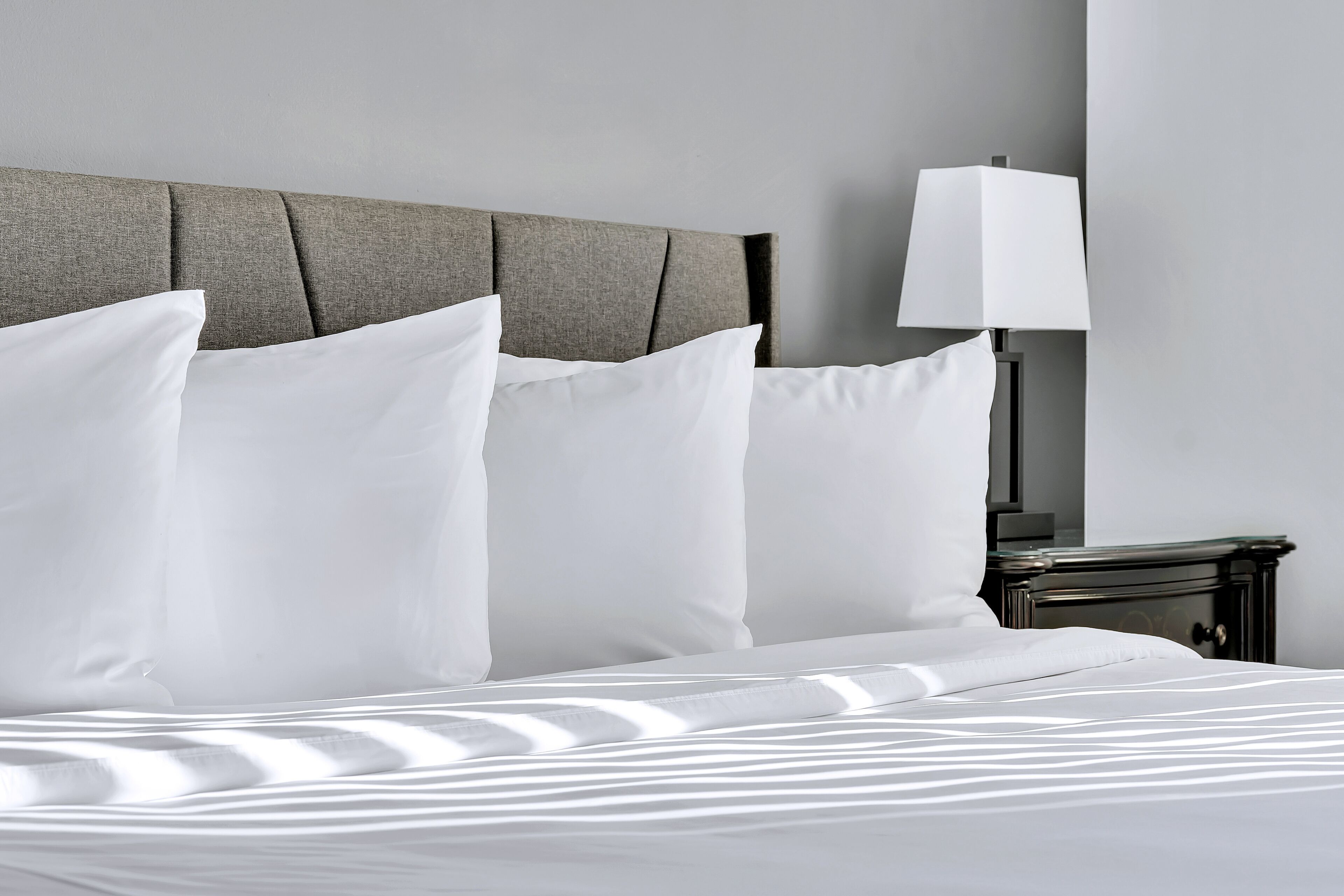 A neatly made bed with white linens and pillows, a grey upholstered headboard, and a bedside table with a lamp.