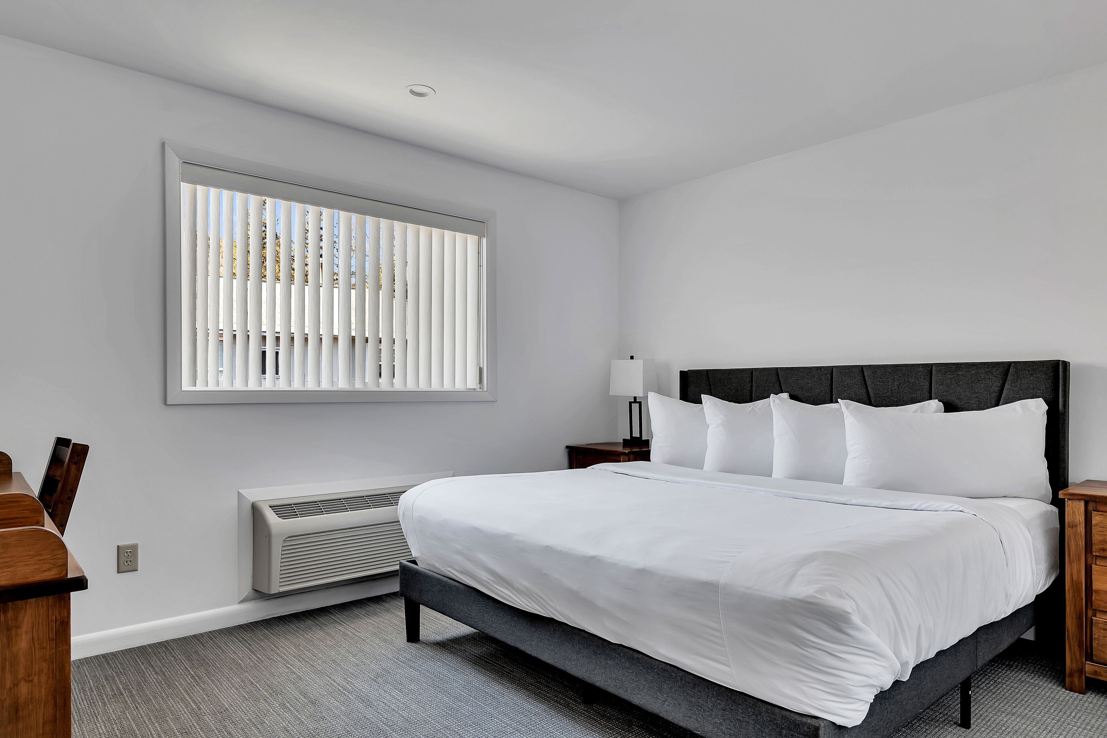 A modern hotel room with a large bed, a bedside table, a lamp, and a window with blinds.