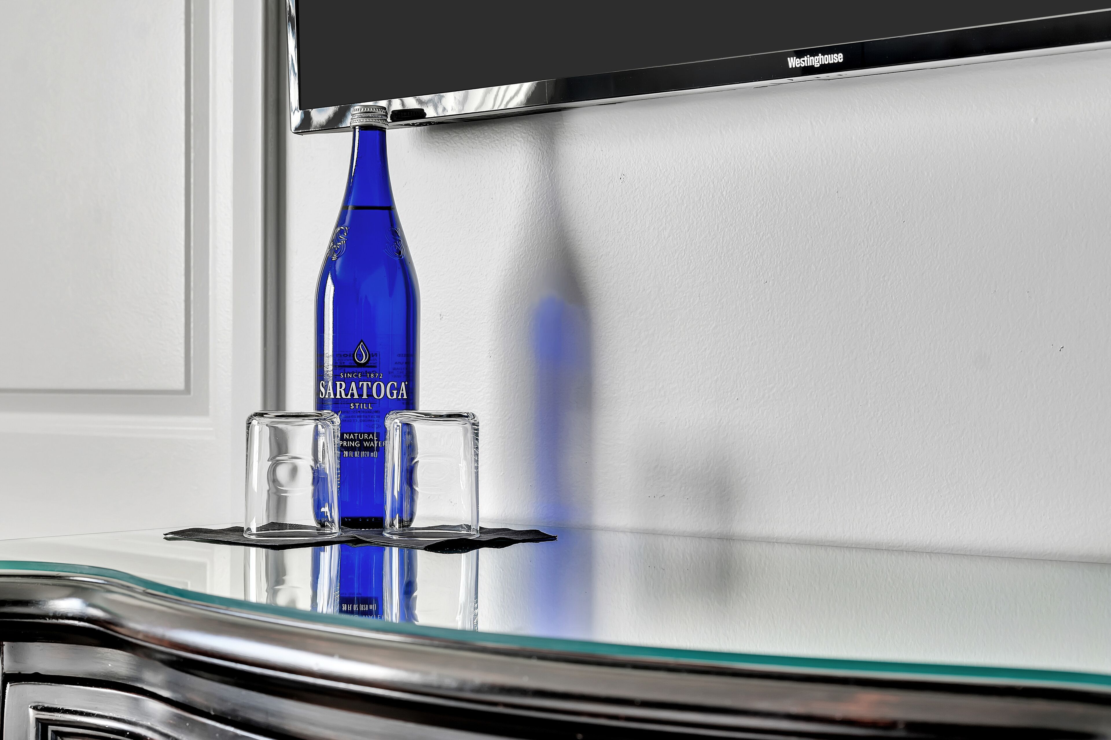 A blue bottle of Sarathoga water, a glass, and a black object on a reflective surface.