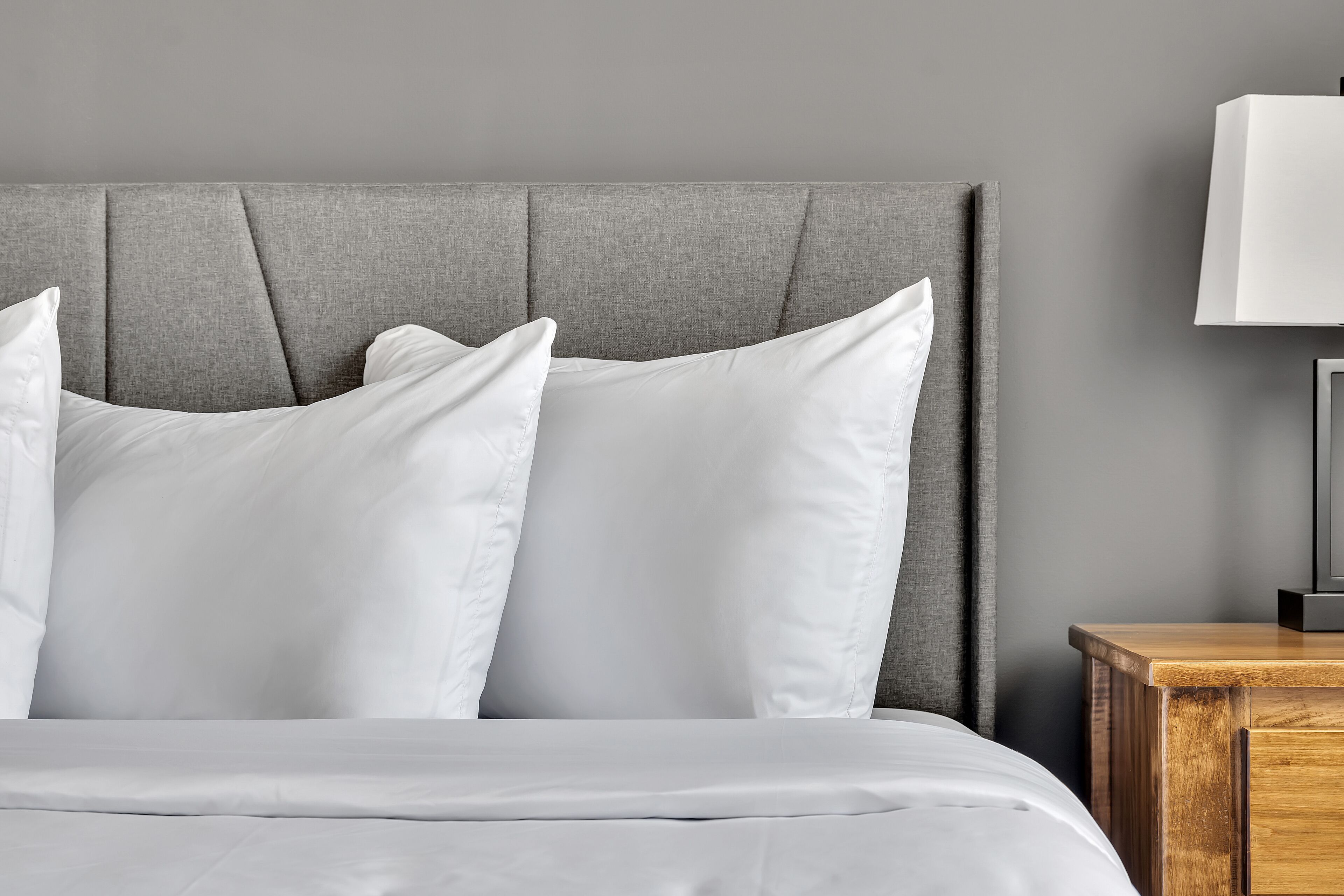Een bed met een grijs gestoffeerd hoofdeinde, wit beddengoed en een houten nachtkastje met een lamp.