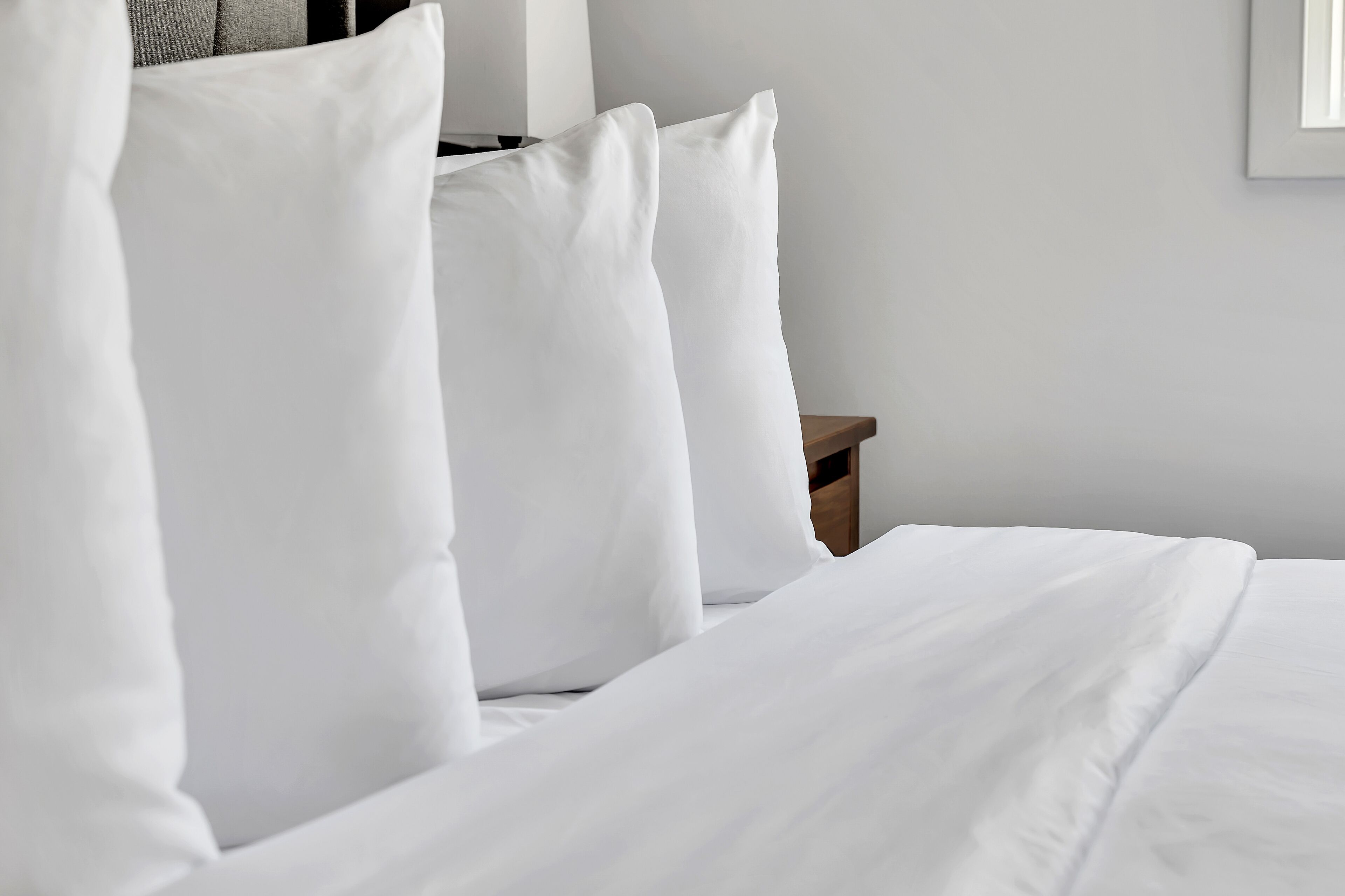 Una cama bien hecha con ropa de cama y almohadas blancas, una mesita de noche de madera y una pared lisa.