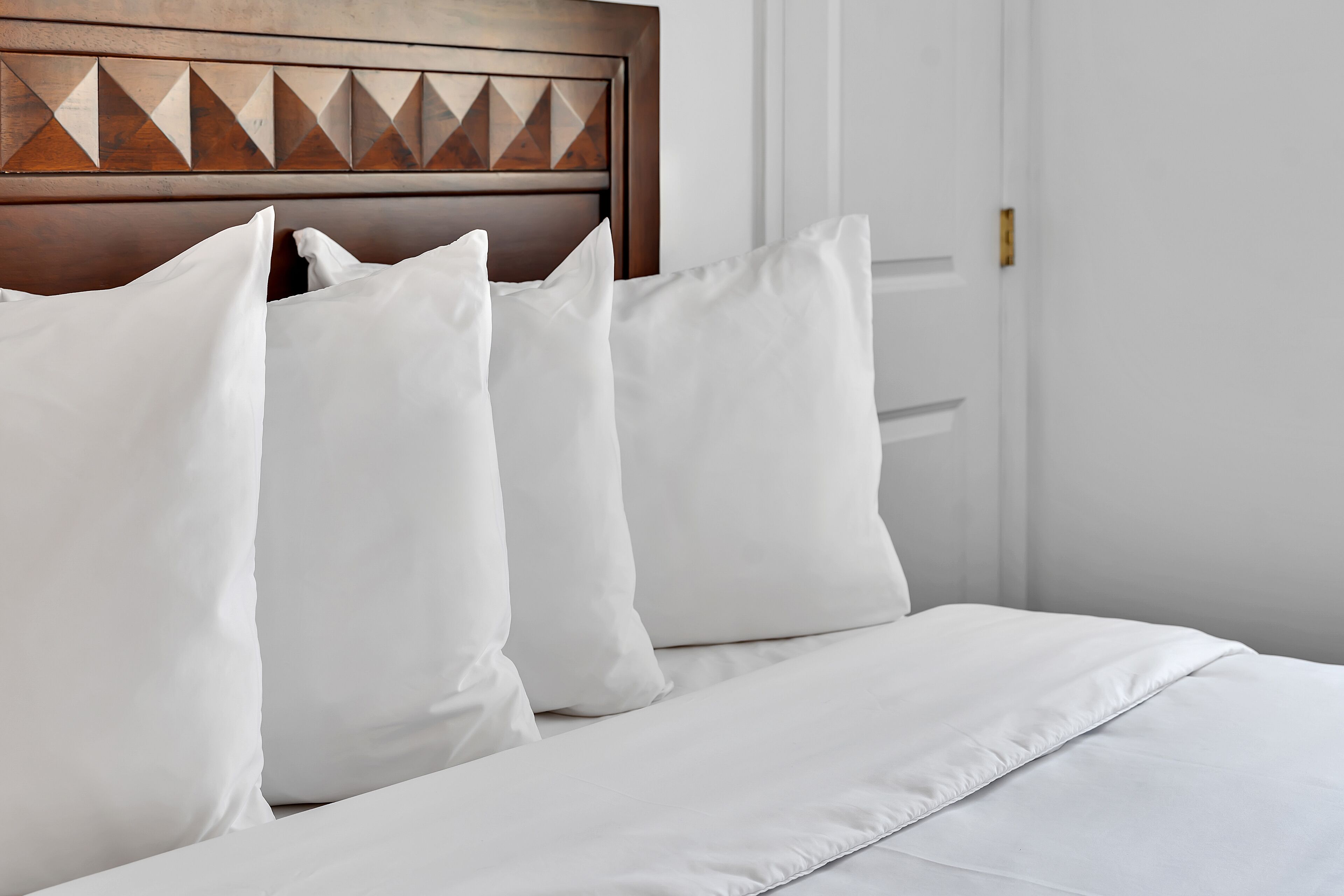 Cama bem arrumada com lençóis e almofadas brancas, cabeceira de madeira com padrão geométrico, e porta branca ao fundo.
