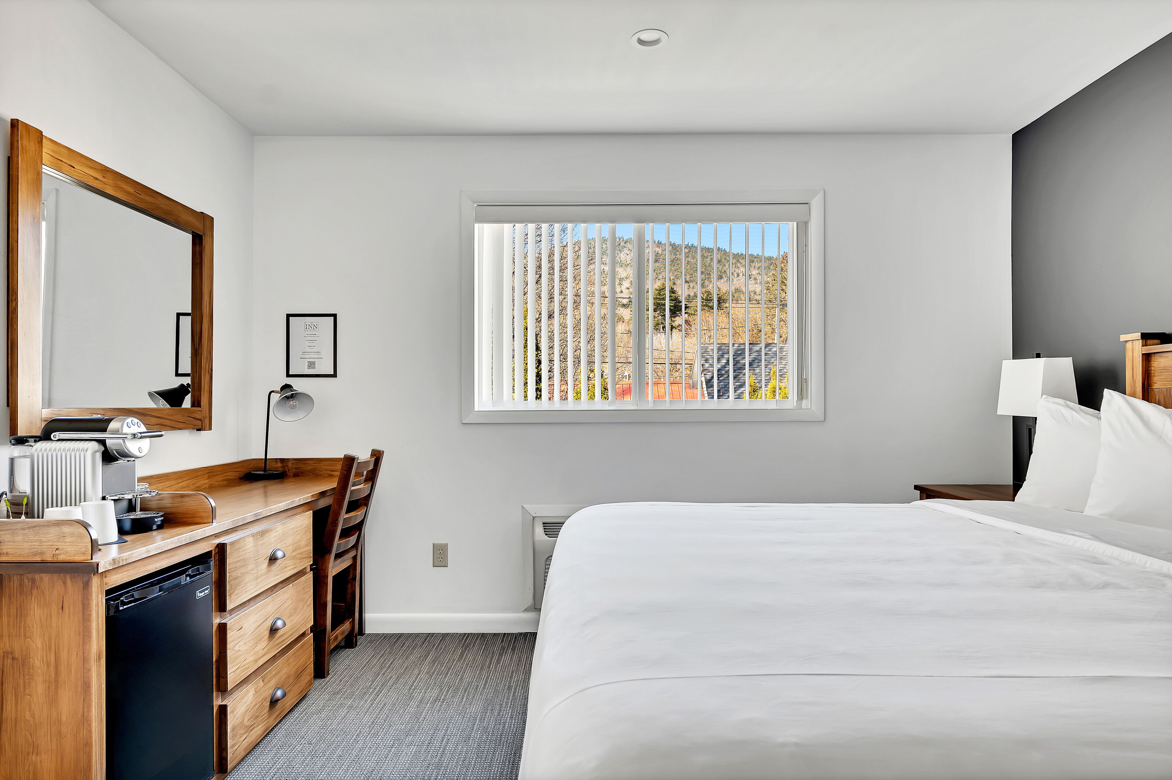 A hotel room with a wooden desk, a bed, a window with blinds, and a radiator.