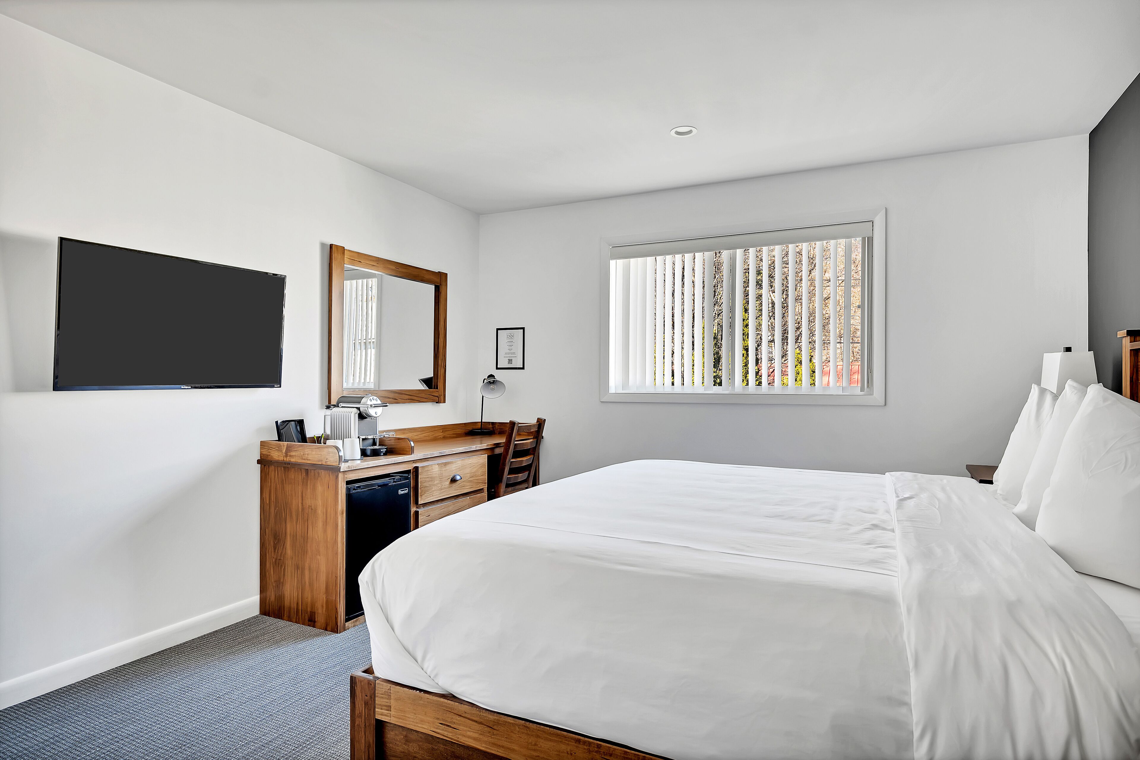 Habitación de hotel con una cama grande, un escritorio con silla, un televisor de pantalla plana montado en la pared y una ventana con persianas verticales.