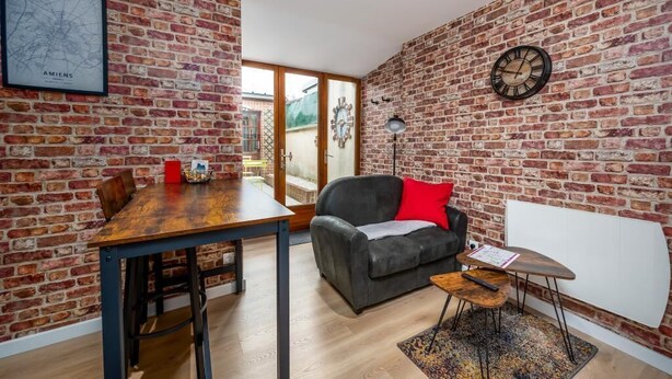 A room with a brick wall, a wooden dining table with chairs, a grey sofa with a red pillow, a small wooden side table, and a clock on the wall. - La parenthèse amiénoise - Cosysejour (Amiens)