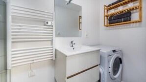 A modern bathroom with a white sink, mirror, and built-in heater. There is a washing machine and a shelf with towels. - La parenthèse amiénoise - Cosysejour (Amiens)