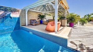 Une piscine dotée d’un espace détente couvert, d’une terrasse en bois et de plantes en pot.