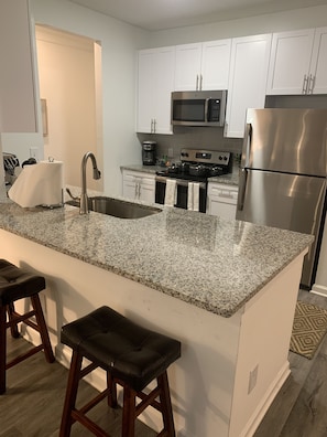 A modern kitchen with white cabinets, a granite countertop, stainless steel appliances, and wooden stools.