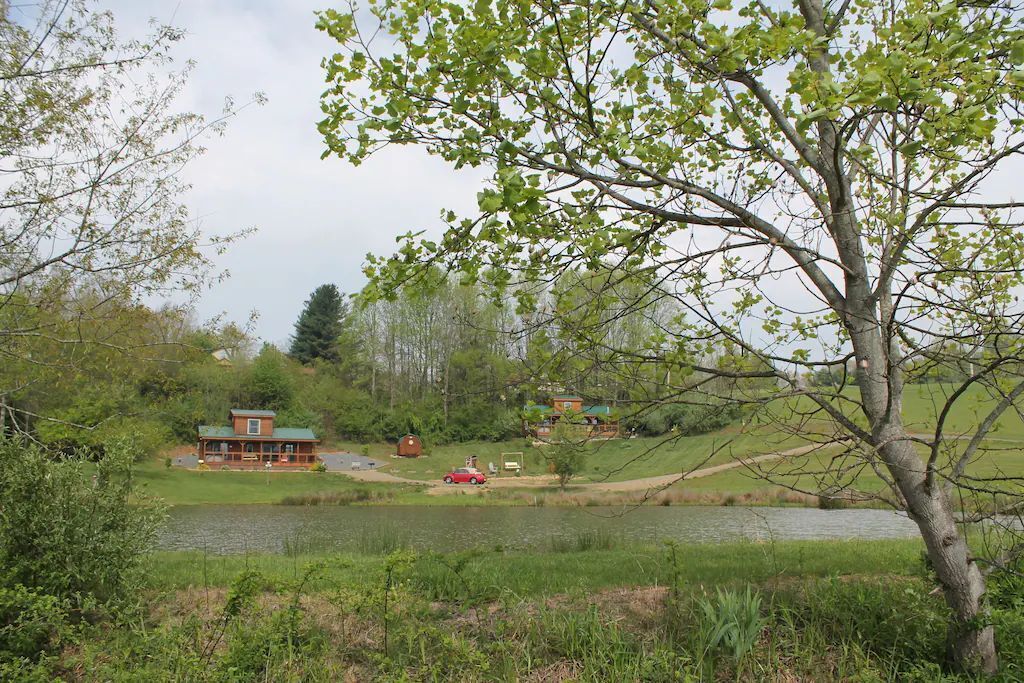 A lakeside cabin with a red car parked in front, surrounded by greenery and a small dock.