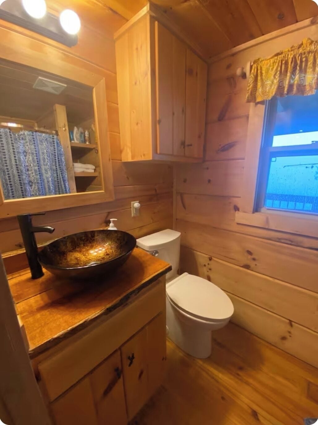 A wooden cabin-style bathroom with a wooden vanity, a large mirror, a sink with a bowl, a toilet, and a window with curtains.