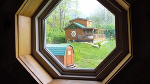 Vista a través de una ventana octogonal de una casa de madera con techo verde, rodeada de un césped cuidado y árboles.