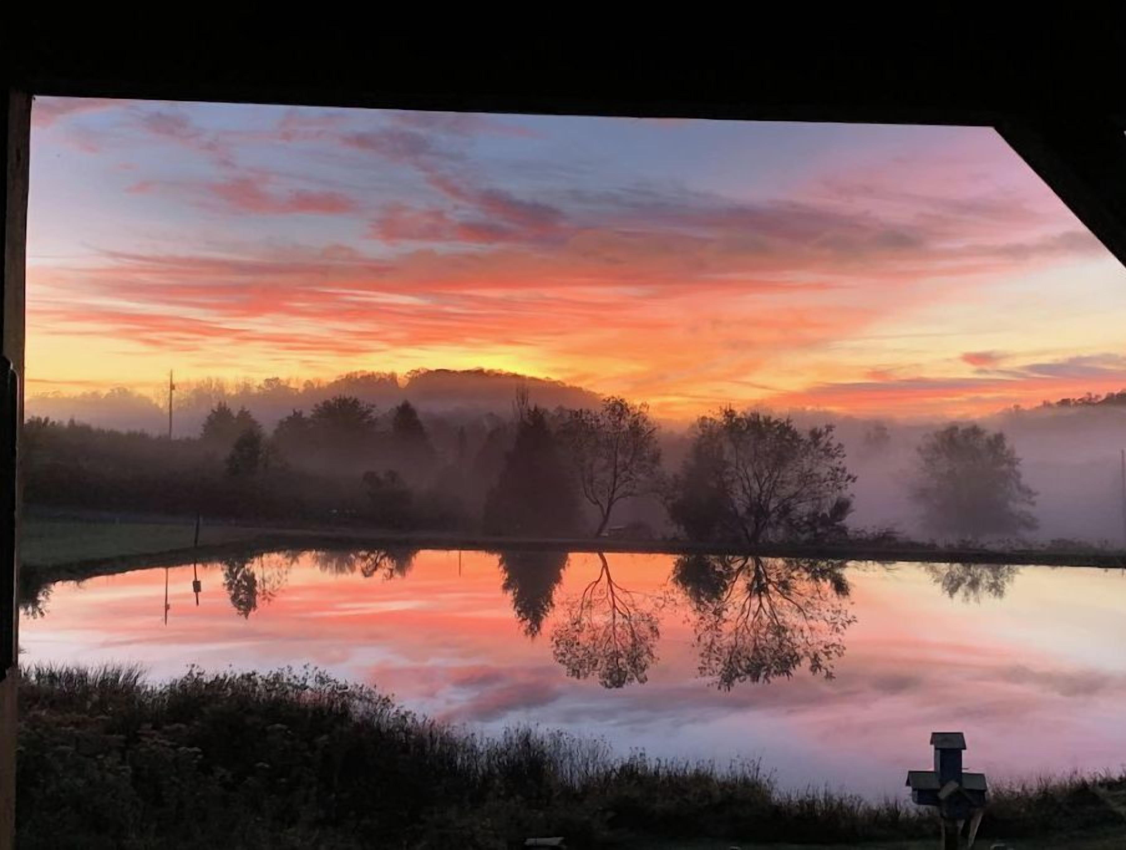 A serene landscape with a calm lake reflecting the sky, surrounded by trees and a misty atmosphere.