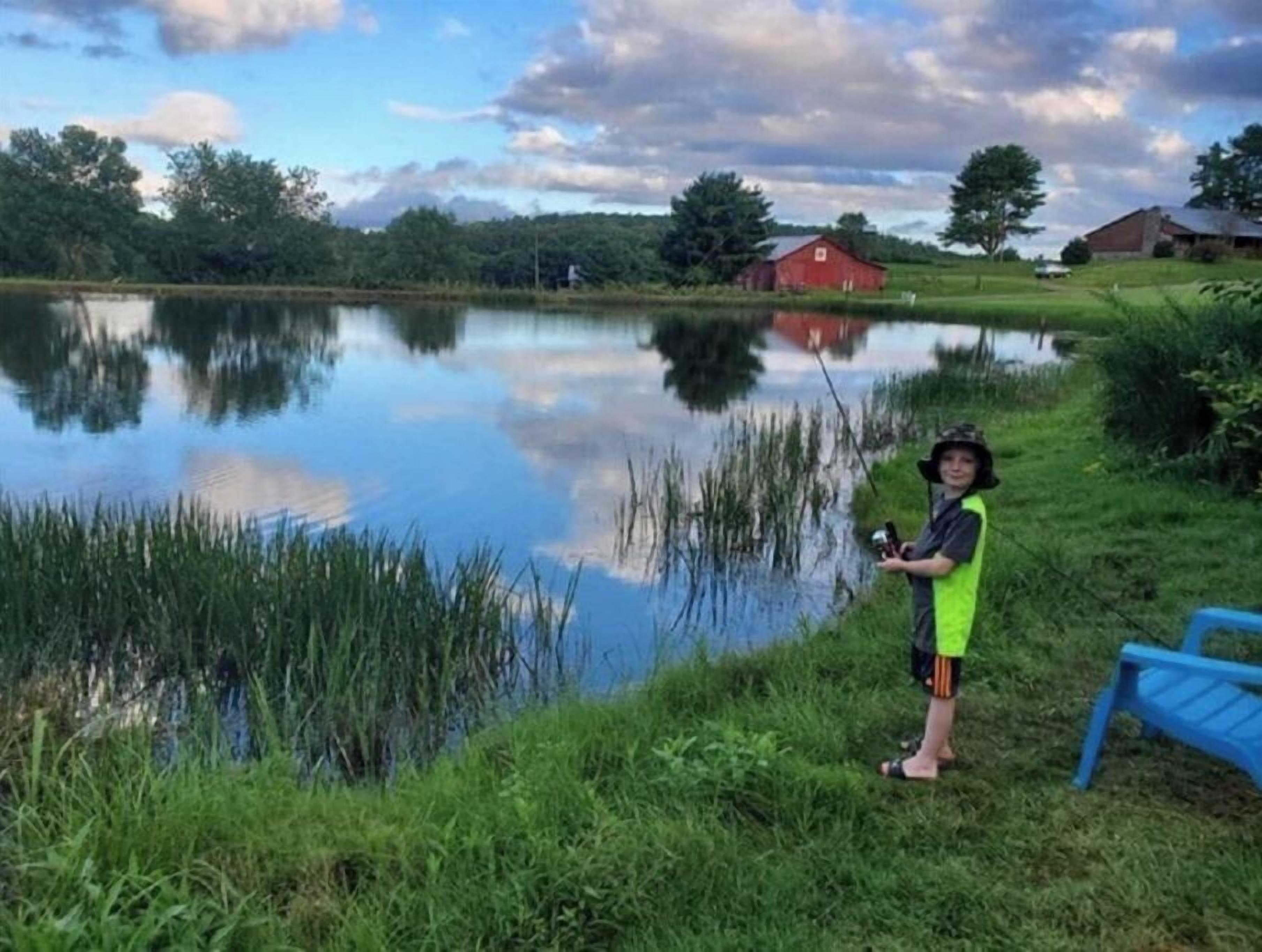 A person fishing by a calm lake with a red barn and houses in the background.