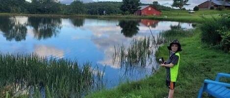 A person fishing by a calm lake with a red barn and houses in the background.