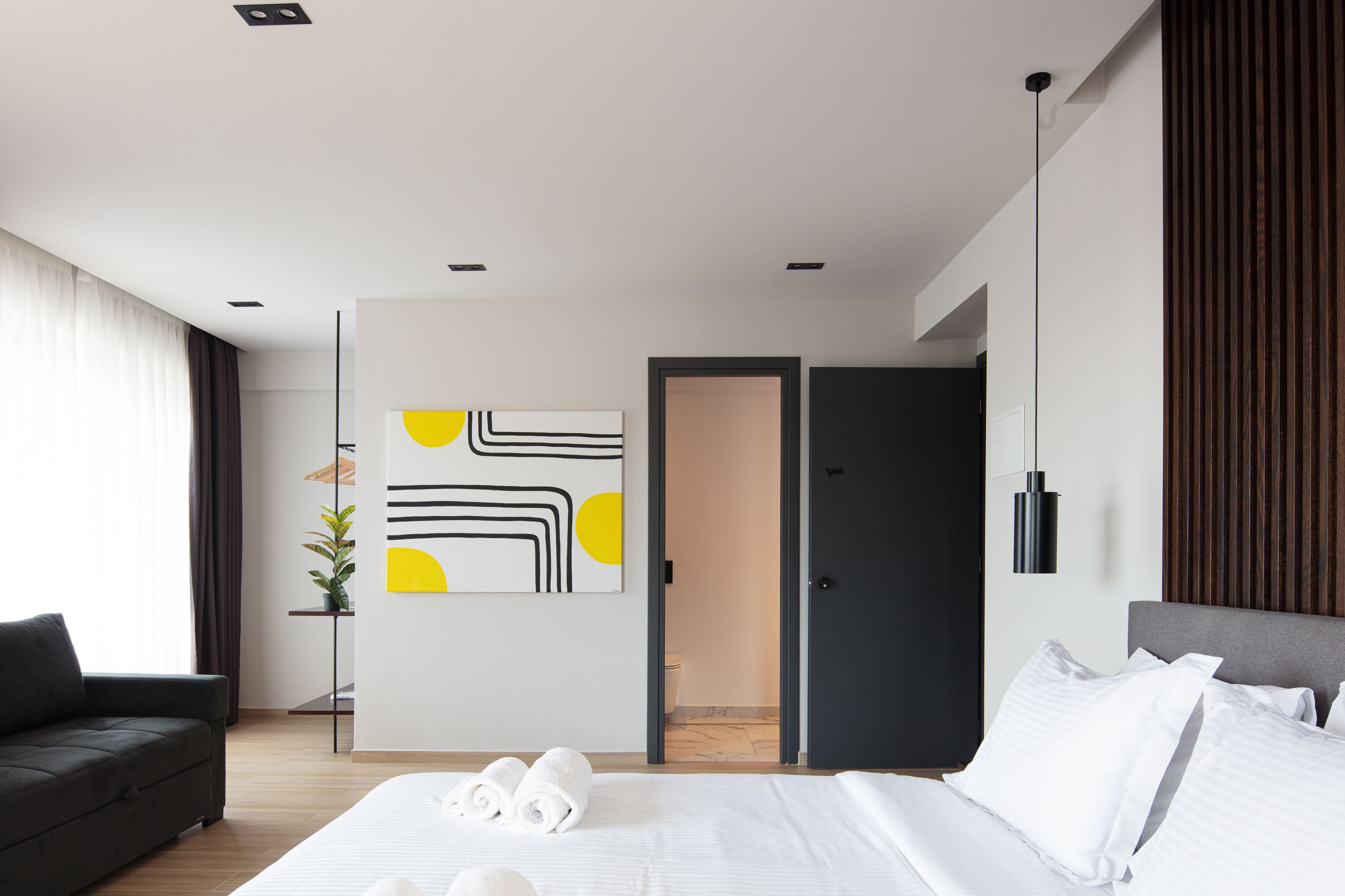 A modern hotel room with a bed, a sofa, a painting, and a door leading to a bathroom.