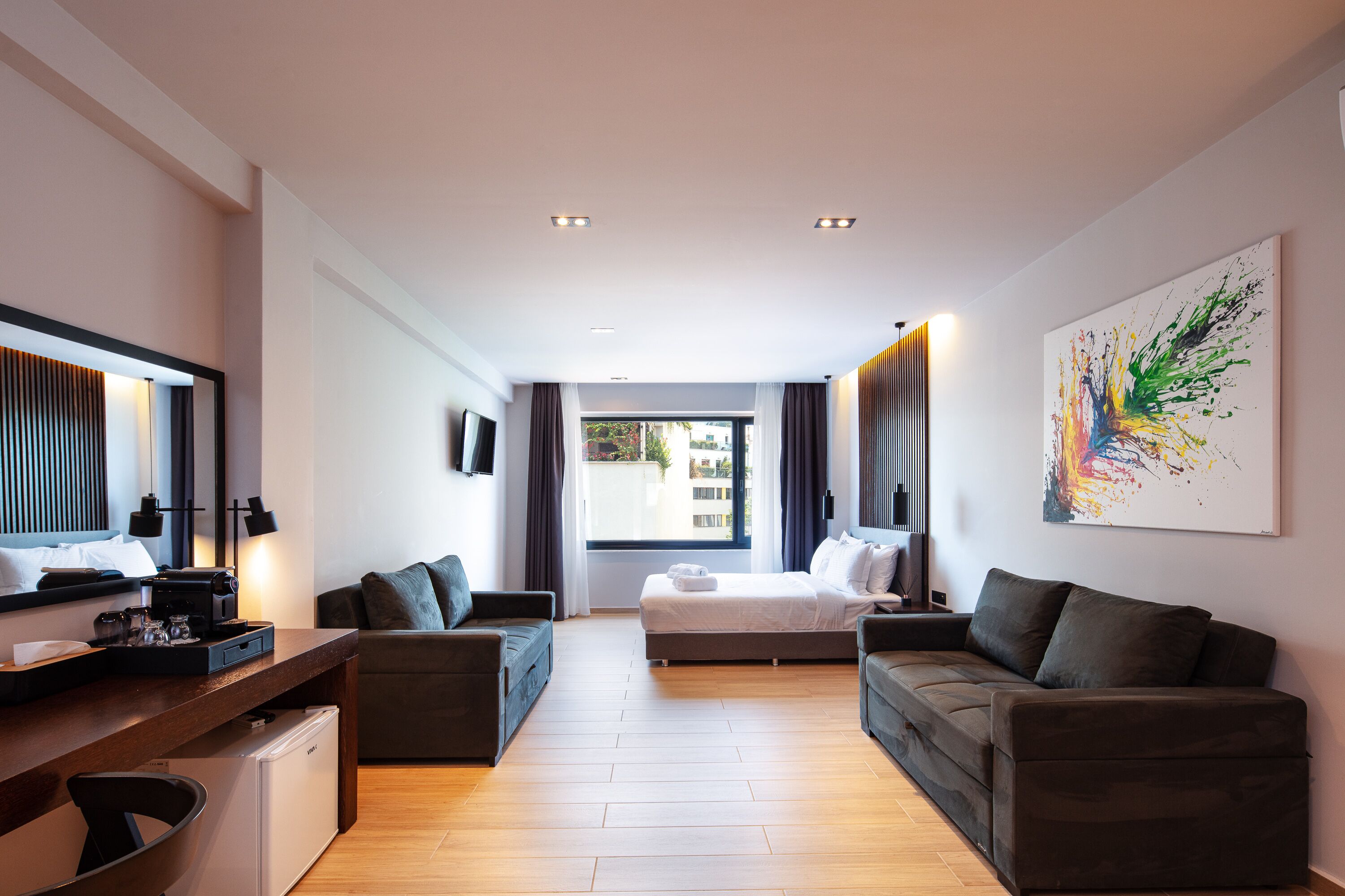 Quarto de hotel moderno com uma cama, dois sofás, uma mesa de trabalho e uma janela grande.
