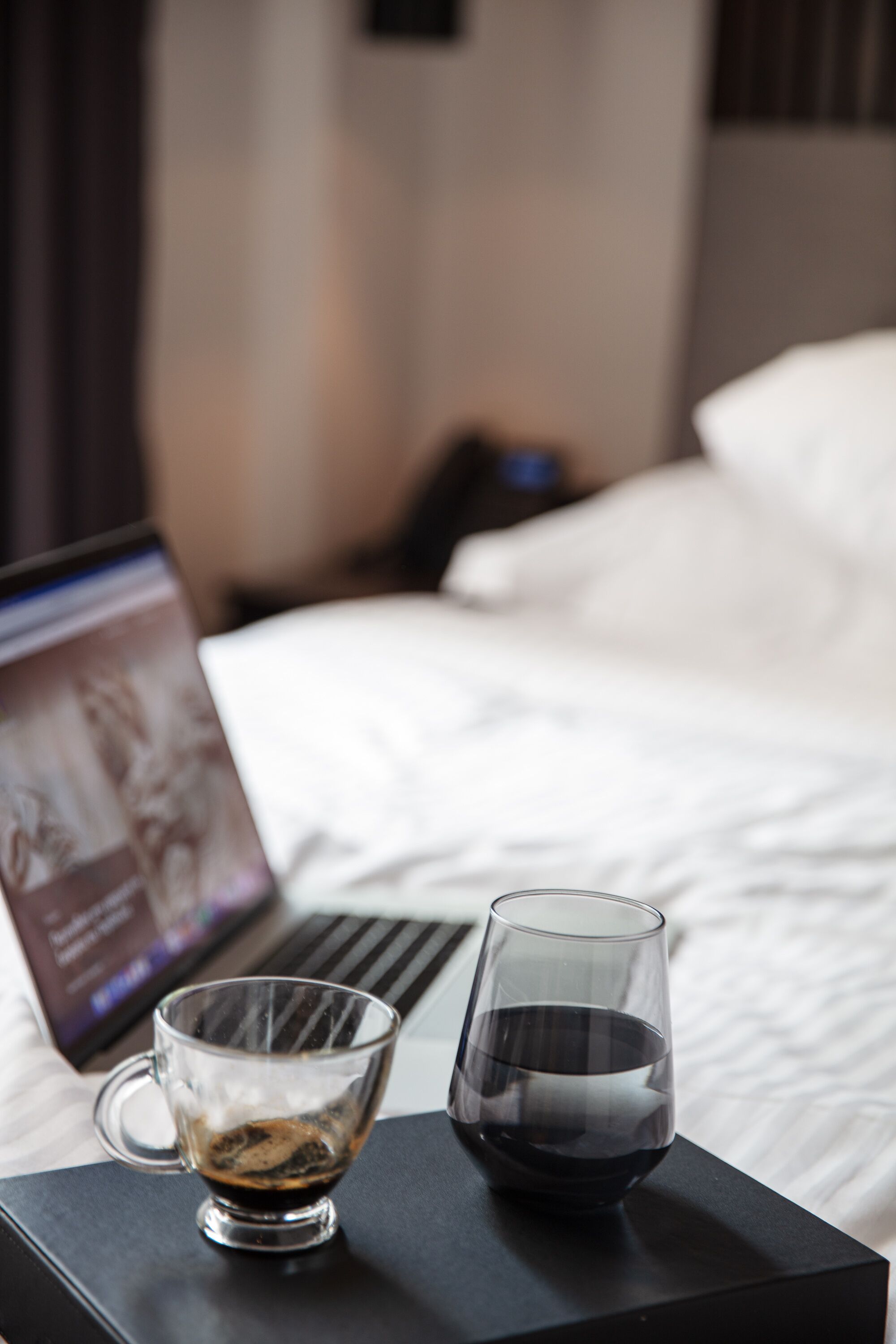 Un ordinateur portable est posé sur une table de chevet, à côté d’un verre contenant un liquide sombre, probablement du café ou du thé, et d’un verre d’eau.